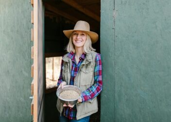Happy female farmer with hat holding feed bowl at ranch door.
