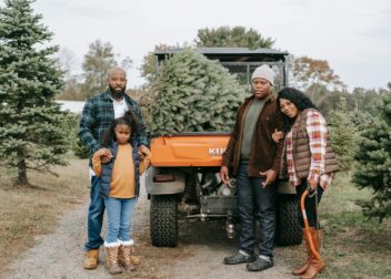 Full body of content African American family looking at camera while standing on farm tree near transport with Christmas tree in trunk