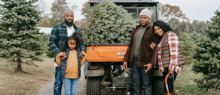 Full body of content African American family looking at camera while standing on farm tree near transport with Christmas tree in trunk